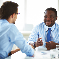 two men in business attire, sitting at a table facing each other, one is facing the camera and the other is facing away