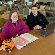 Holly sits at a library table with her computer and book receiving assistance from a College Navigator
