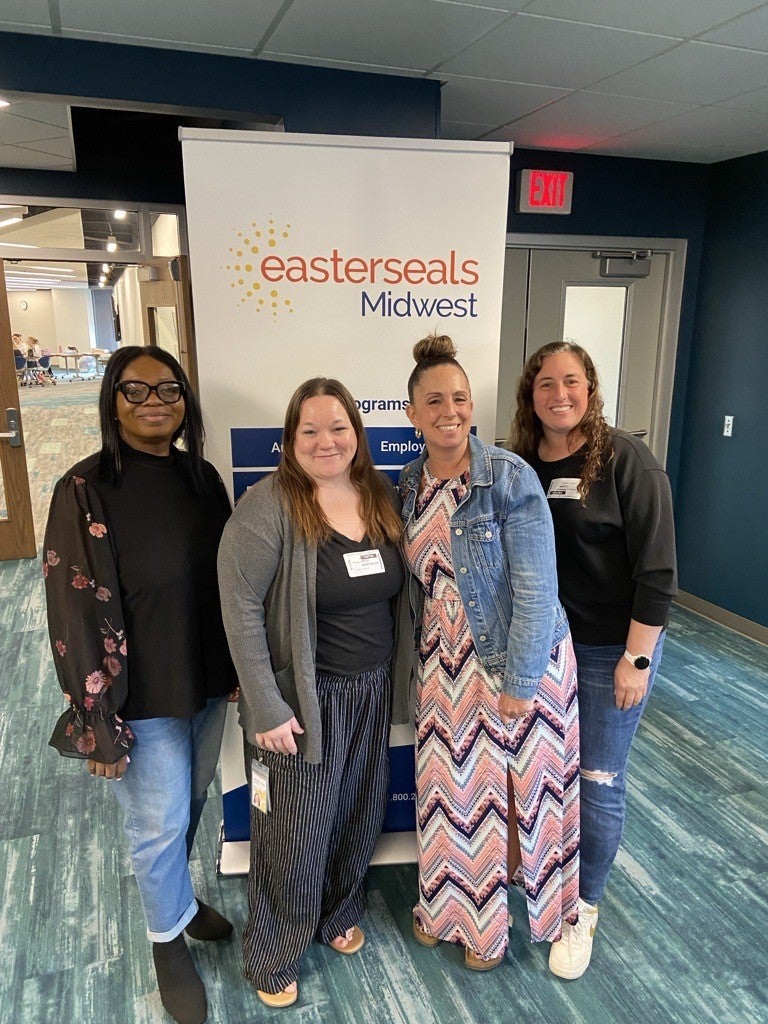 Four women smile and pose in front of an "Easterseals Midwest" banner inside a building with teal-colored flooring. They are wearing casual to business-casual outfits, including jeans, cardigans, a floral blouse, and a long patterned dress with a denim jacket. The atmosphere appears friendly and welcoming, likely at a work-related event or gathering.