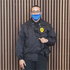 Elijah M. is wearing his new work uniform: a security guard uniform. He is also wearing a blue mask and glasses. He is posing in front of a planked fence, holding his radio.
