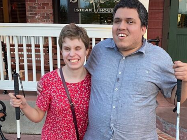 Alicia and her boyfriend, both using a white cane, are outside and smiling 