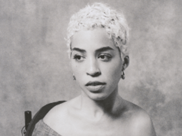 Jillian Mercado in an over-the-shoulder blouse, short hair, gazing to the side