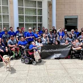 A group of people with disabilities in front of a building, holding a banner that says "#TeachDisabilityHistory"