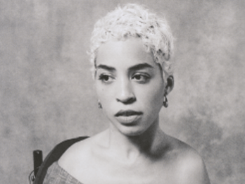Jillian Mercado in an over-the-shoulder blouse, short hair, gazing to the side