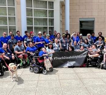 A group of people with disabilities in front of a building, holding a banner that says "#TeachDisabilityHistory"